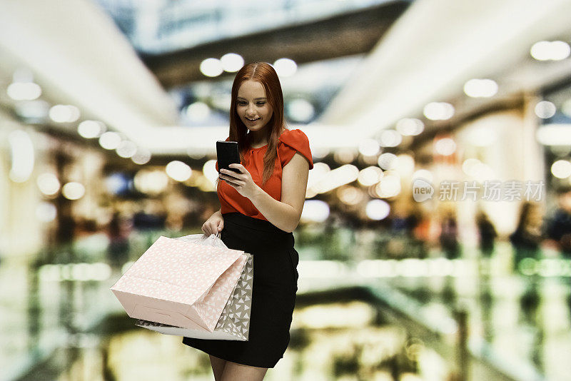 正面视图18-19岁的红发年轻女性购物狂/十几岁的女孩站在商店/购物中心/百货公司/服装店前的模糊背景谁是微笑的/快乐的/愉快的，使用智能手机/销售